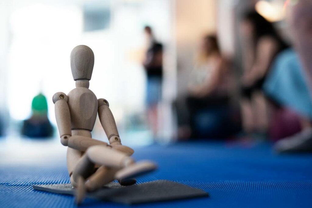 Muñeco articulado de madera en postura sentada sobre una colchoneta azul en un centro de pilates, con personas practicando en el fondo desenfocado.